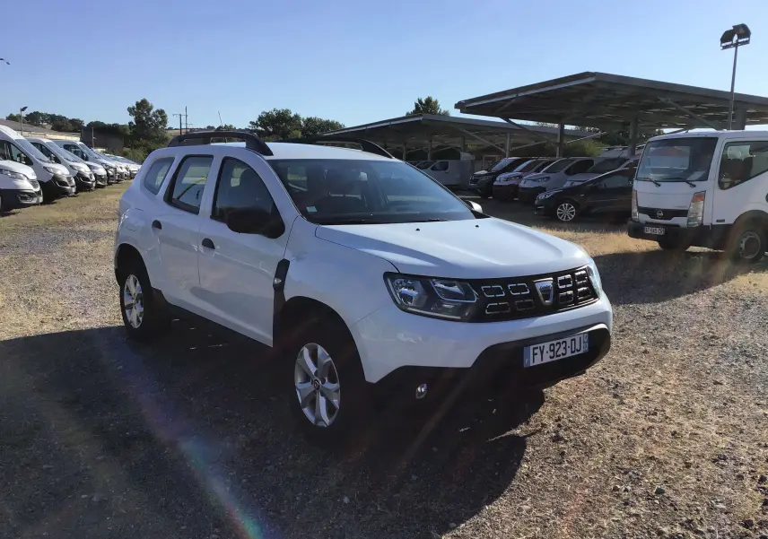 Dacia Duster blanc vu en 3/4 avant droit sur un terrain avec plusieurs véhicules en arrière-plan.