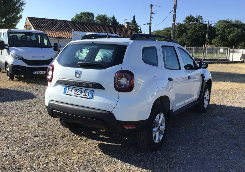 Vue 3/4 arrière droite d'un Dacia Duster blanc 2021 avec barres de toit et feux arrière LED sur un terrain extérieur.