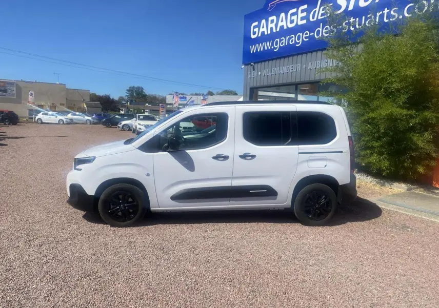Profil droit d'un Citroën Berlingo blanc 2025 avec portes coulissantes et jantes noires, stationné devant un garage.