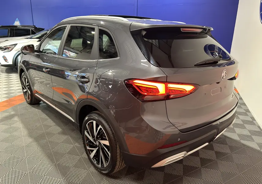 Vue 3/4 arrière droite du SUV MG ZS Hybrid+ 197 Luxury en gris Candem avec feux arrière allumés et toit panoramique.