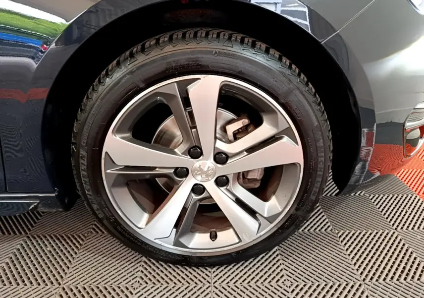 Gros plan sur la roue avant droite d'une Peugeot 308 gris foncé, mettant en valeur la jante alliage et le logo lion central.