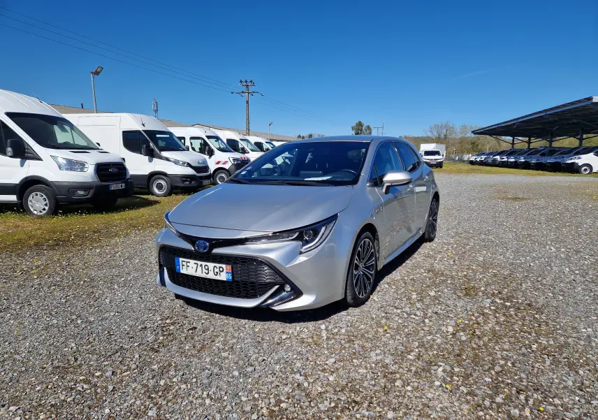 Toyota Corolla hybride gris clair métal vue 3/4 avant droit sur un parking ensoleillé avec plusieurs utilitaires blancs en arrière-plan.