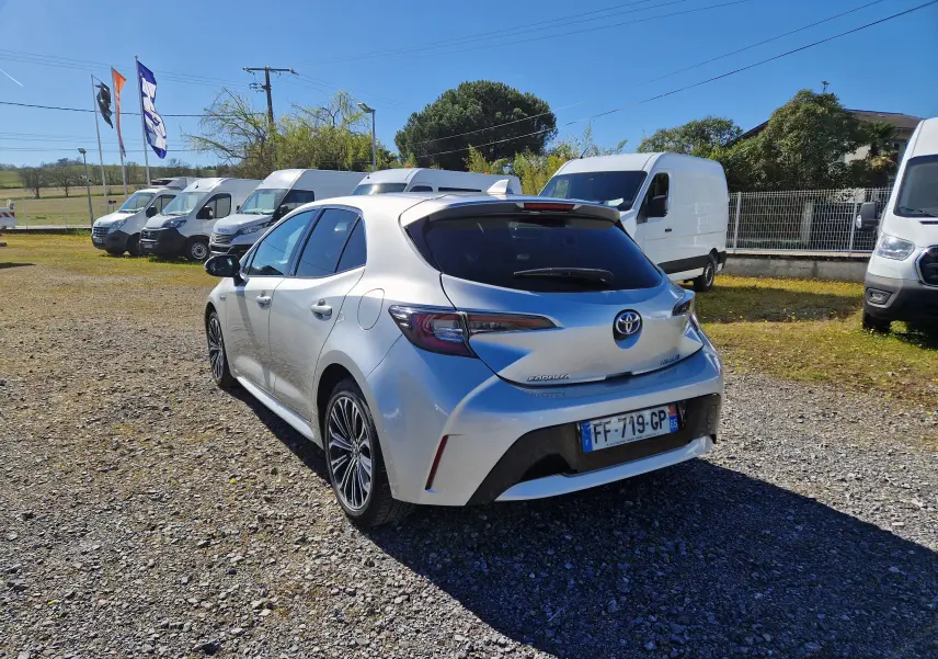 Toyota Corolla hybride gris clair métal vue 3/4 arrière droit sur parking avec plusieurs fourgons blancs en arrière-plan.