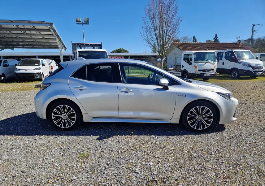 Vue de profil côté gauche d'une Toyota Corolla hybride gris clair métal avec jantes alliage et badge hybride visible.