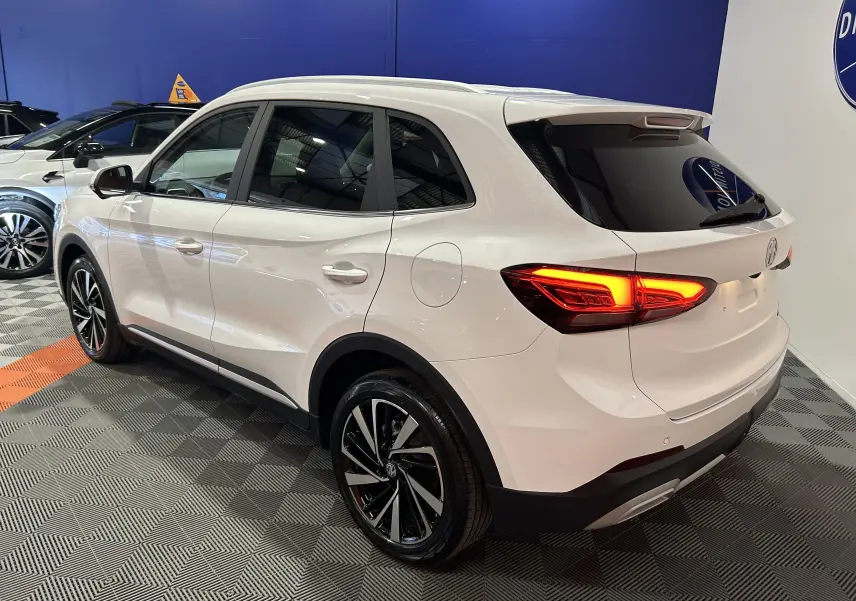 Vue 3/4 arrière droite du MG ZS Hybrid+ 197 Luxury blanc Dover avec toit panoramique ouvrant en intérieur showroom.