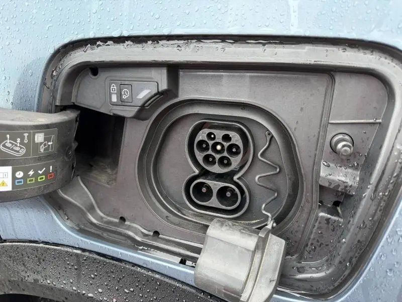 Prise de recharge électrique ouverte sur le côté gauche d’un Peugeot 3008 GT bleu 2024 sous la pluie.