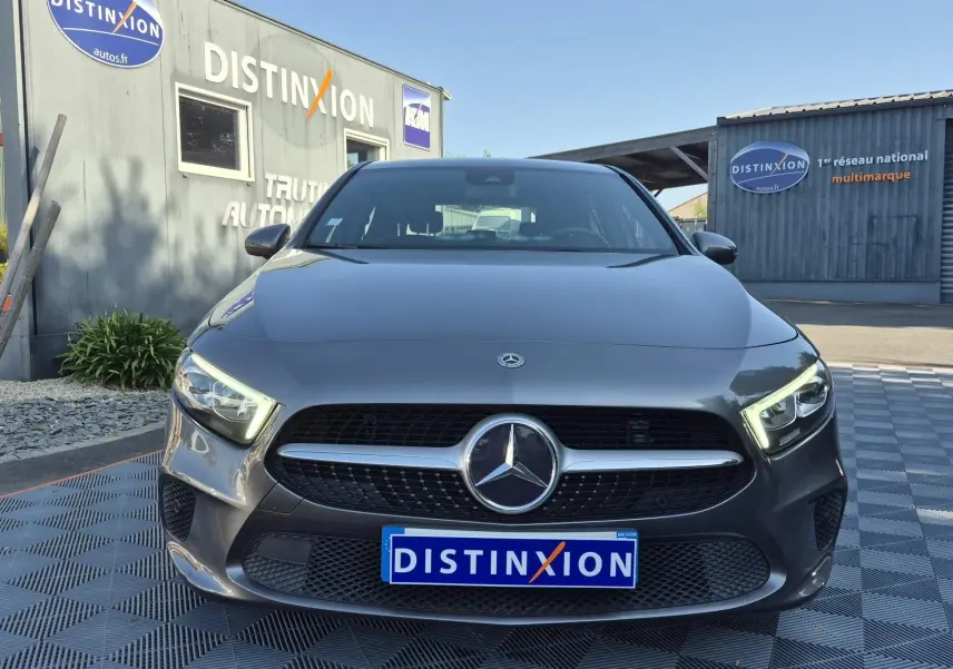 Vue frontale d'une Mercedes Classe A 180d gris foncé avec calandre noire et phares LED allumés, devant un bâtiment Distinxion.
