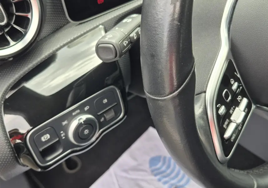Vue rapprochée de l’intérieur côté gauche du volant cuir noir d’une Mercedes Classe A 180d gris foncé, avec commandes éclairage.