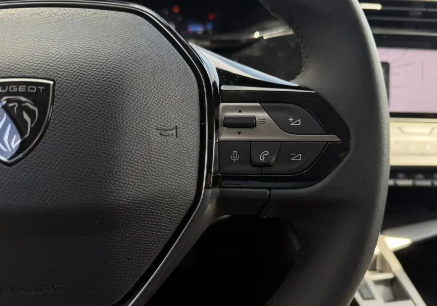 Gros plan sur le volant cuir noir du Peugeot 308 SW 2025 avec commandes audio et logo lion au centre.
