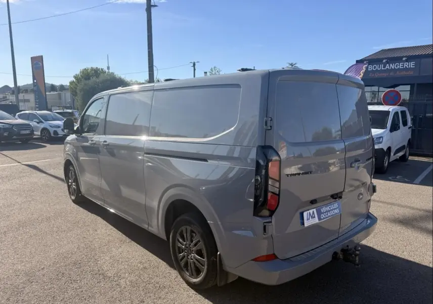 Vue 3/4 arrière droite d’un Ford Transit Custom Fourgon gris 2024 avec hayon et attelage visible.
