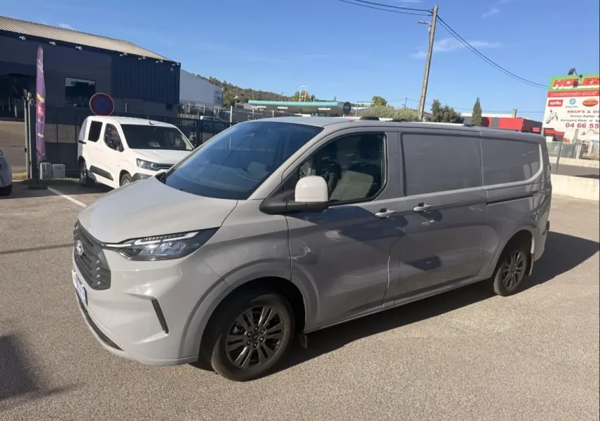 Ford Transit Custom Fourgon gris, vue 3/4 avant droit, avec jantes foncées et calandre marquée, stationné en extérieur.