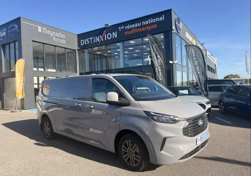 Ford Transit Custom Fourgon gris en 3/4 avant droit devant un showroom multimarque par temps clair.