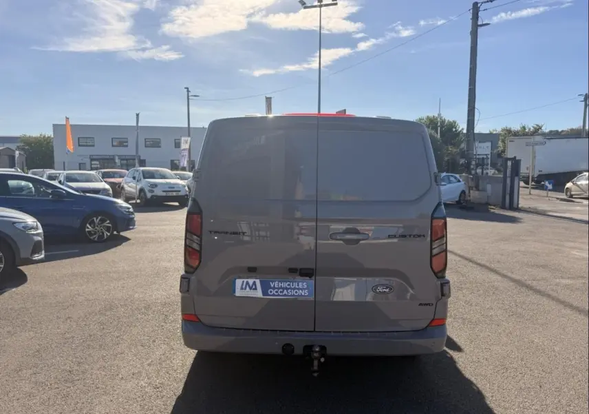 Vue arrière d’un Ford Transit Custom gris 2024 avec hayon fermé et attelage visible sur parking extérieur.