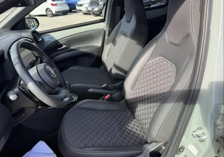 Vue intérieure côté conducteur de la Toyota Aygo X 2025, sièges noirs avec surpiqûres losangées et volant multifonction.