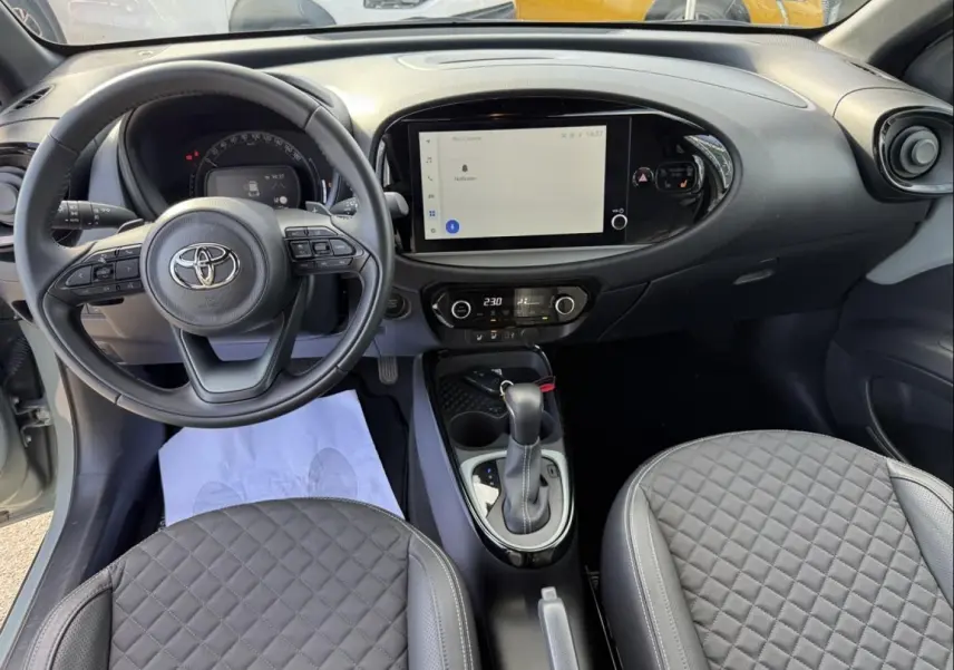 Vue intérieure frontale du tableau de bord et volant de la Toyota Aygo X 2025, avec écran tactile et sellerie grise matelassée.