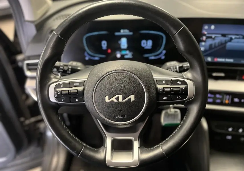 Volant cuir noir avec commandes intégrées et logo KIA, tableau de bord numérique flou en arrière-plan du Kia Sportage 2022.