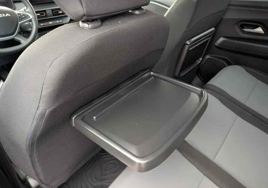 Vue intérieure arrière droite du Dacia Jogger 2025 avec tablette rabattable sur siège avant et sellerie gris clair et noir.