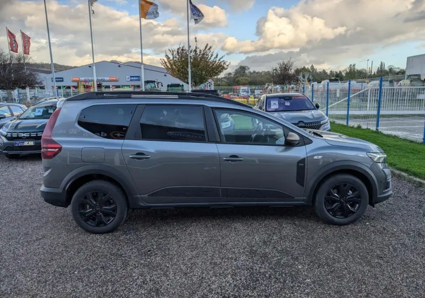 Vue de profil côté gauche d'un Dacia Jogger gris métallisé avec jantes noires et barres de toit sur parking extérieur.