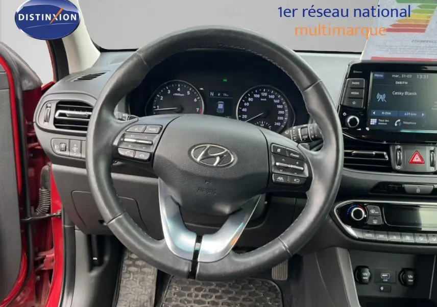 Vue intérieure centrée sur le volant cuir de la Hyundai i30 SW 2023 rouge Sunset Red Metal avec tableau de bord et écran tactile.