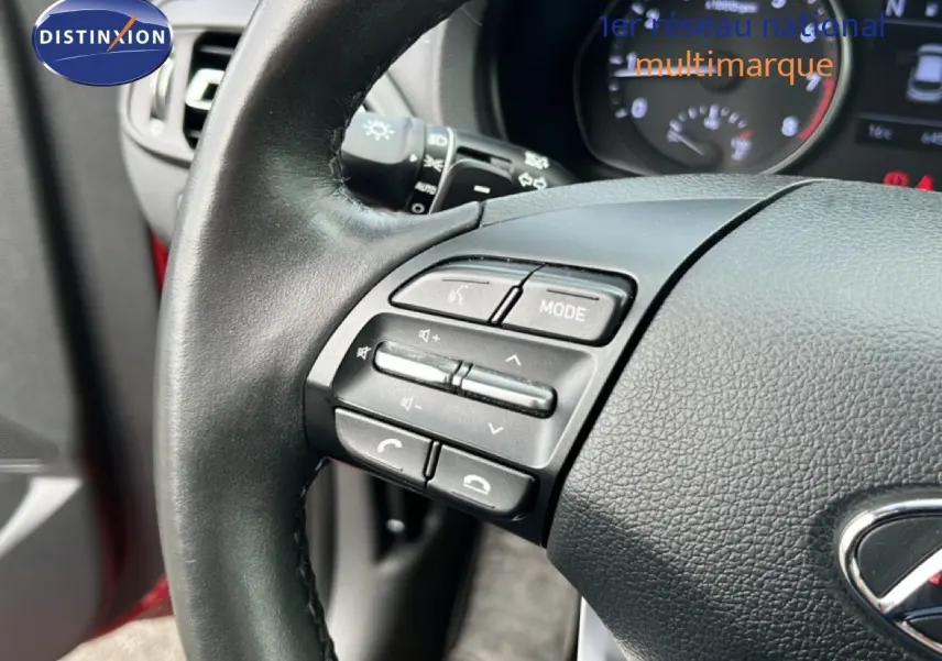 Gros plan sur le volant côté gauche de la Hyundai i30 SW 2023 rouge, avec commandes audio et tableau de bord en arrière-plan.