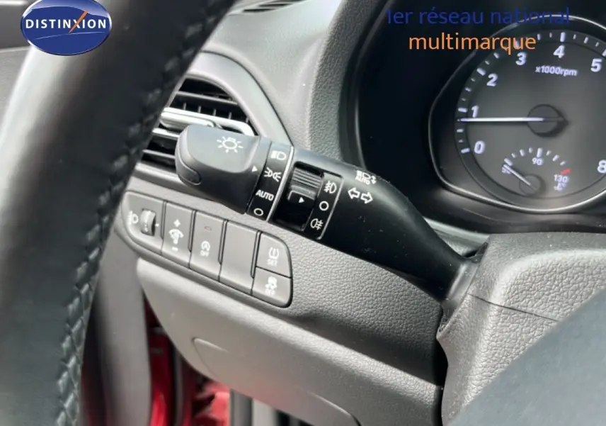 Gros plan sur la commande des feux et les boutons de réglage à gauche du volant dans l’habitacle d’une Hyundai i30 SW rouge.