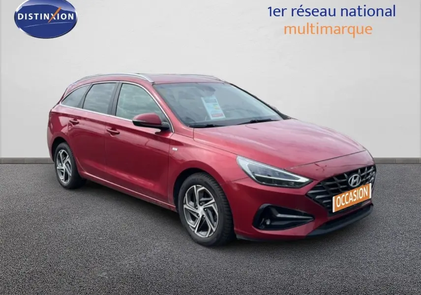 Vue 3/4 avant gauche d'une Hyundai i30 SW 2023 rouge Sunset Red Metal avec jantes bi-ton et calandre noire.