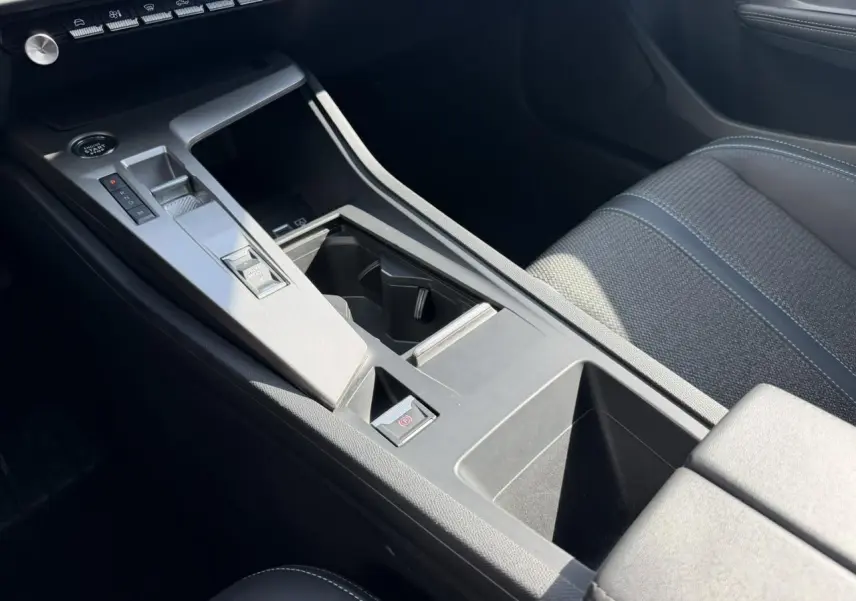 Vue intérieure rapprochée de la console centrale et accoudoir avant de la Peugeot 308 SW 2025, sièges tissu gris avec surpiqûres bleues.