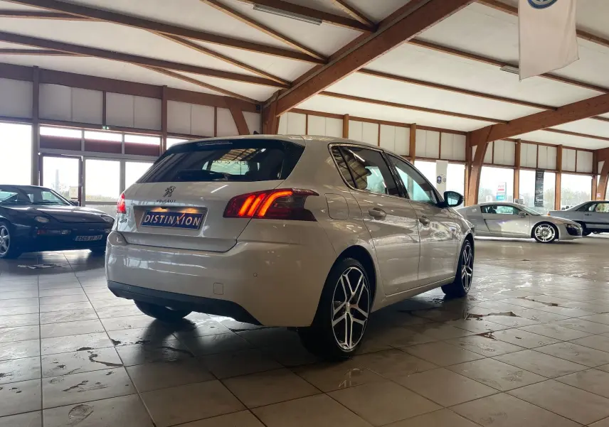 Vue 3/4 arrière droite d'une Peugeot 308 blanche 5 portes avec feux arrière allumés dans un showroom.