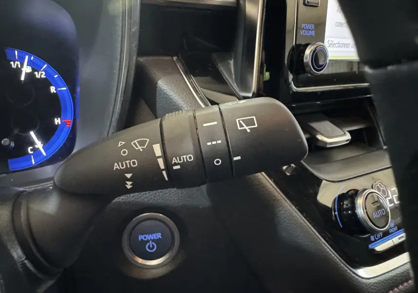 Gros plan sur la commande d’essuie-glace et le bouton Power bleu du tableau de bord d’une Toyota Corolla hybride gris clair.