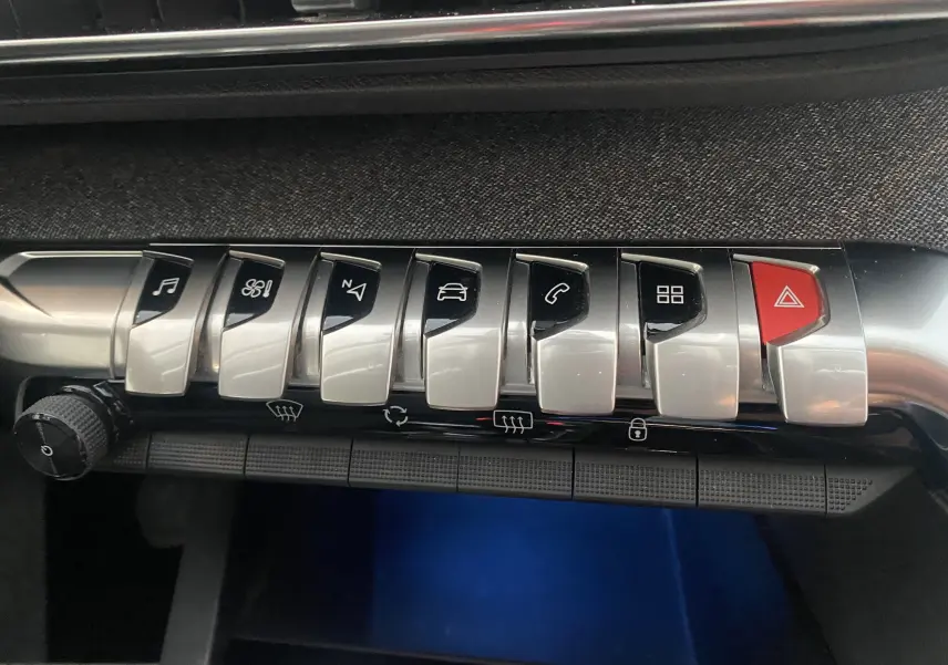 Gros plan sur la rangée de commandes chromées du tableau de bord du Peugeot 5008 blanc, avec bouton rouge d'alerte.