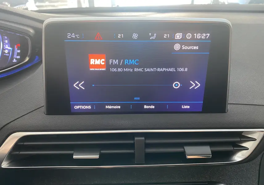 Écran multimédia central du Peugeot 5008 blanc, affichant la radio RMC avec commandes tactiles et aérateurs dessous.