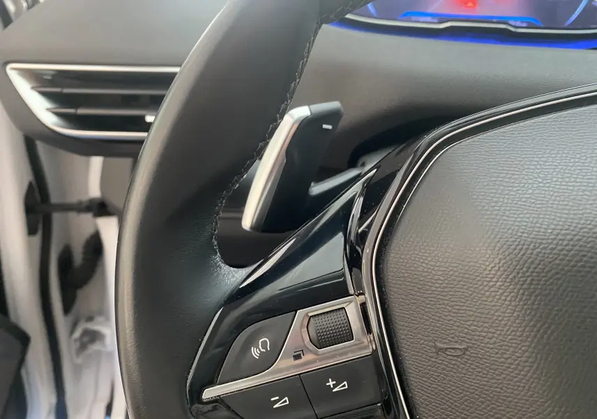 Gros plan sur la commande gauche du volant en cuir noir du Peugeot 5008 blanc, avec palettes de changement de vitesse visibles.