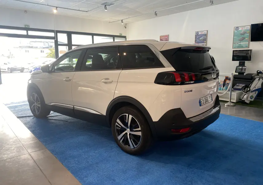 Peugeot 5008 blanc blanquise vu de trois quarts arrière droit en intérieur, avec attelage neuf et jantes modernes.