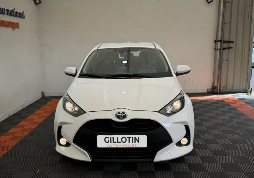 Vue frontale d'une Toyota Yaris hybride blanche 2022 avec phares allumés dans un garage showroom.