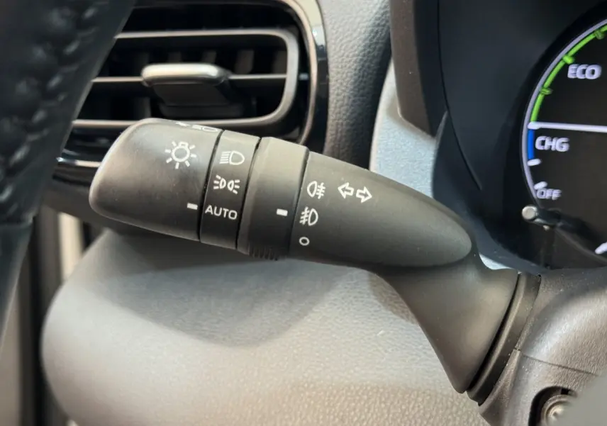 Gros plan sur la commande des phares et clignotants à gauche du volant dans l'habitacle noir de la Toyota Yaris hybride blanche.