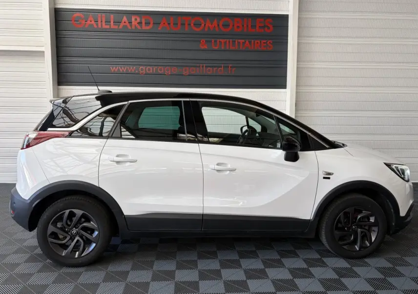 Vue de profil côté gauche d'un Opel Crossland blanc avec toit noir et jantes foncées, stationné en intérieur.