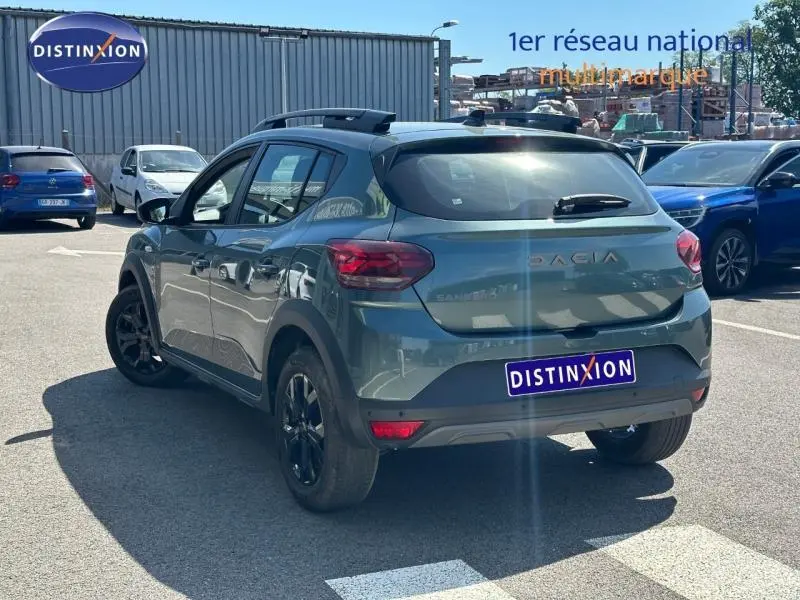 Vue 3/4 arrière droite d'une Dacia Sandero Stepway 2025 vert cèdre métallisé avec barres de toit et jantes noires.