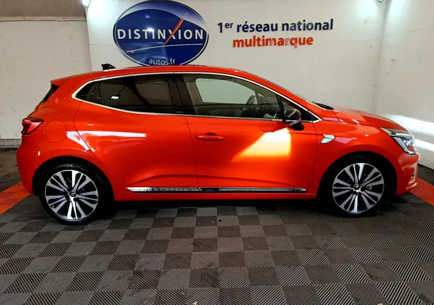Profil droit d'une Renault Clio Initiale Paris E-TECH 140 orange, avec jantes alliage et vitres teintées, en intérieur showroom.