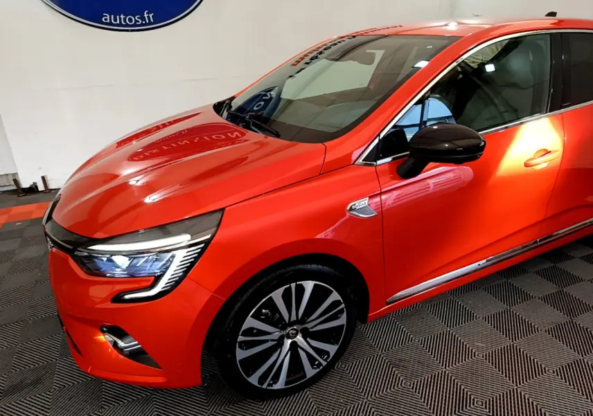 Renault Clio Initiale Paris orange vue 3/4 avant droit avec phares LED allumés et jantes distinctives.