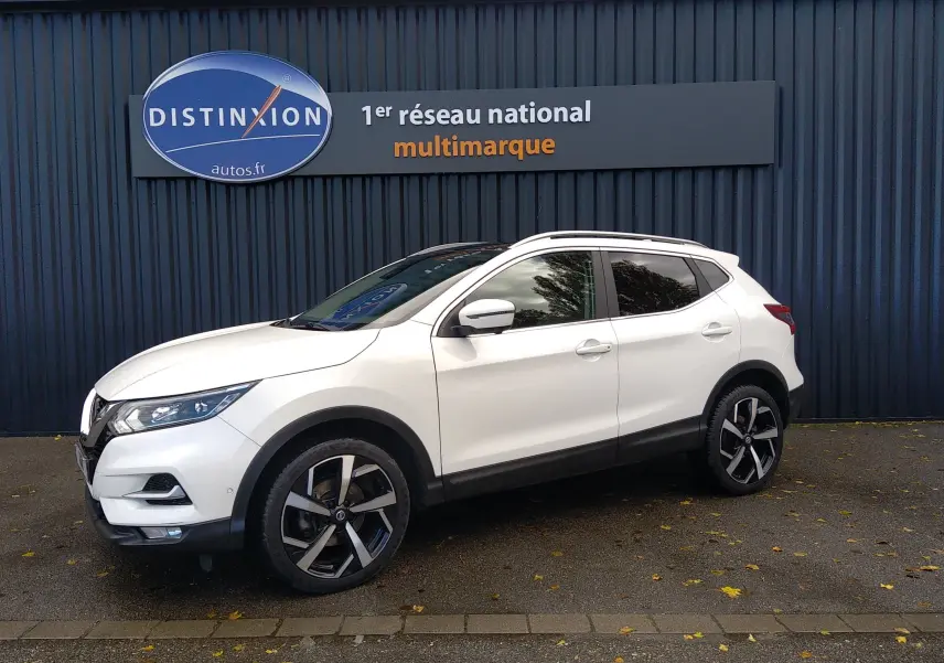 Nissan Qashqai blanc en 3/4 avant droit, avec jantes alliage noires et toit ouvrant, devant un mur sombre.