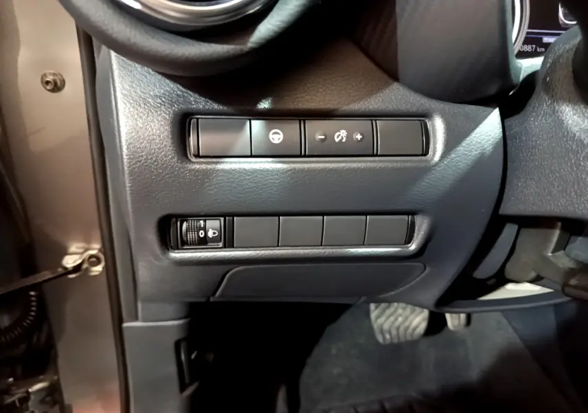 Vue rapprochée des commandes de réglage d’éclairage et assistance au volant côté gauche du Nissan Juke Hybrid 2023 gris foncé.