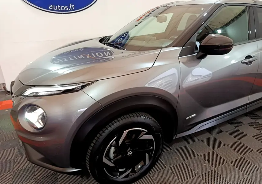 Nissan Juke Hybrid 143 Business+ gris foncé vu en 3/4 avant droit avec jantes noires et phares LED allumés.