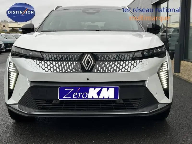 Vue frontale d'une Renault Scénic E-Tech électrique blanche avec calandre à motifs hexagonaux et feux LED verticaux allumés.