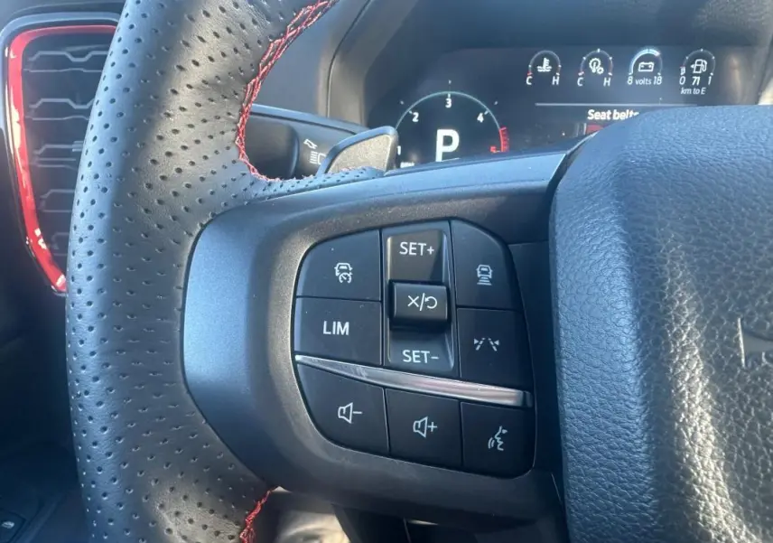 Gros plan sur les commandes à gauche du volant perforé noir avec surpiqûres rouges du Ford Ranger blanc, tableau de bord numérique en arrière-plan.