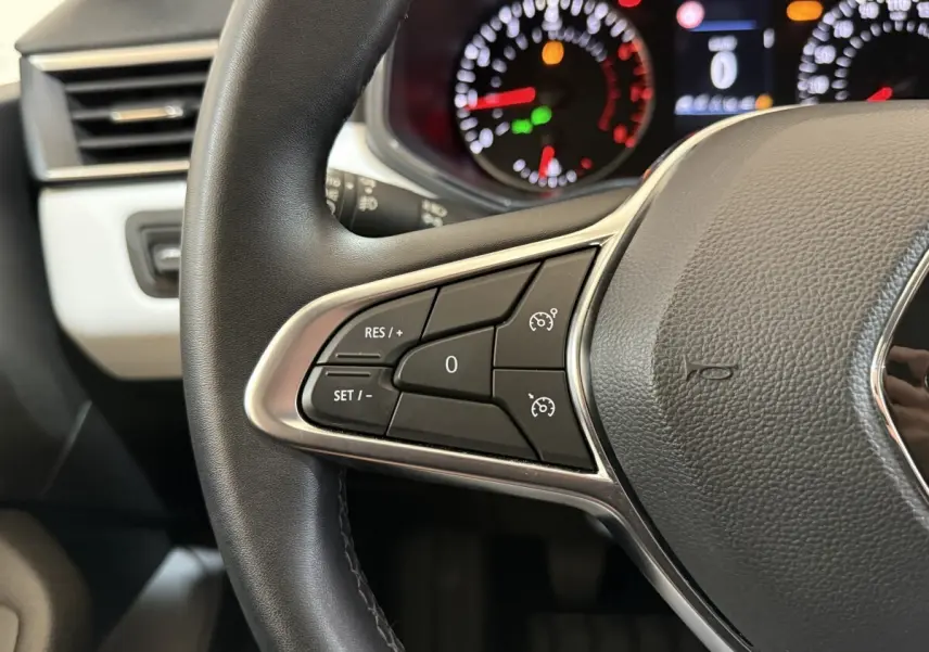 Gros plan sur le volant cuir synthétique de la Renault Clio blanc, avec commandes de régulateur de vitesse et tableau de bord flou.