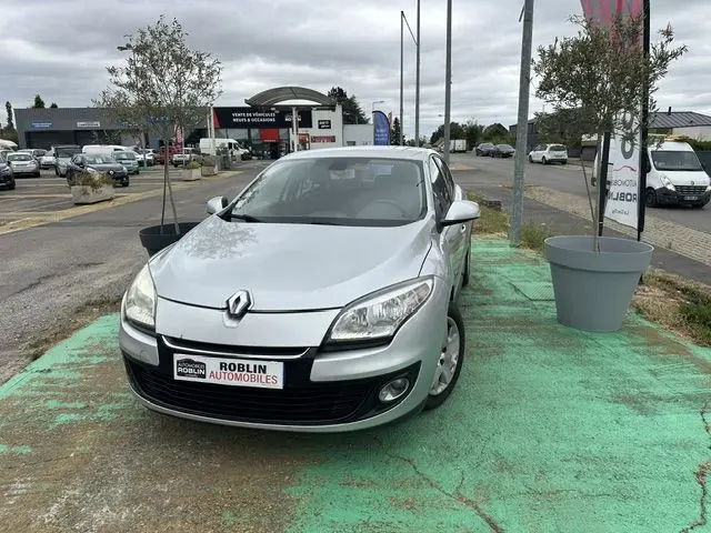 Renault Mégane 3 gris vue de face, légèrement décalée côté gauche, sur parking extérieur avec panneaux publicitaires.