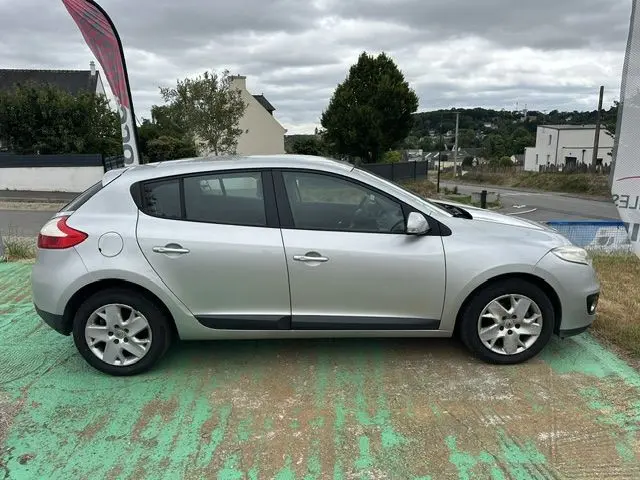 Profil côté gauche d'une Renault Mégane 3 gris clair, cinq portes, stationnée sur sol vert usé, sous ciel nuageux.