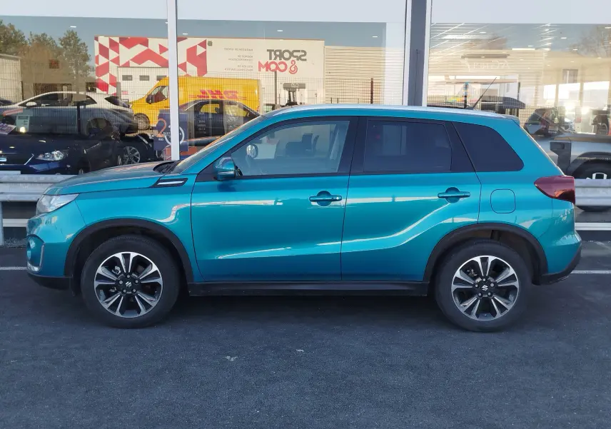 Suzuki Vitara 1.5 Hybrid 2023 en turquoise métallisé, vue de profil côté gauche sur parking devant vitrine.