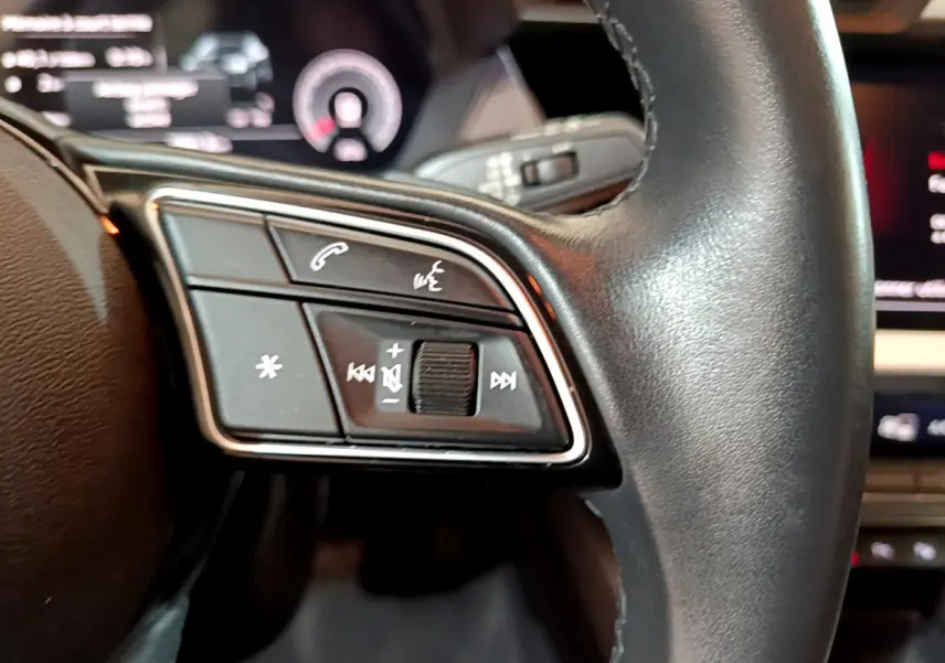 Gros plan sur les commandes gauche du volant cuir noir de l'Audi A3 35 TFSI 2023 avec affichage numérique flou en arrière-plan.