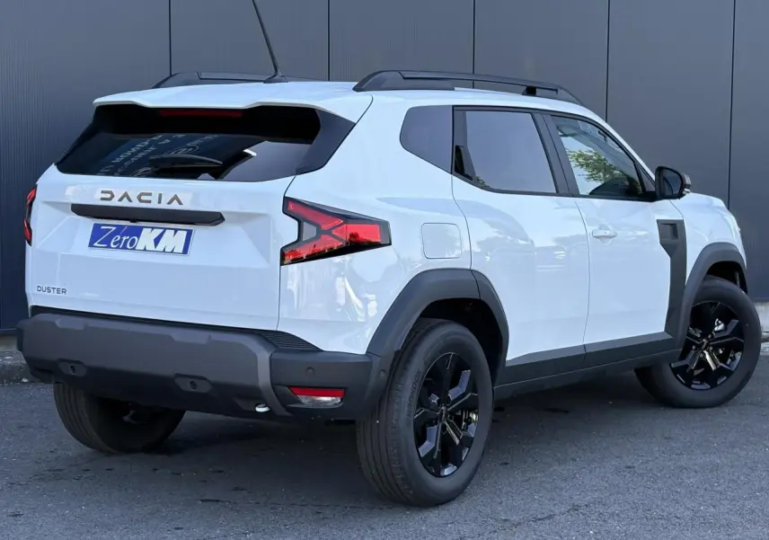 Vue 3/4 arrière droite du Dacia Duster 2026 blanc Glacier avec jantes alliage noires et barres de toit noires.